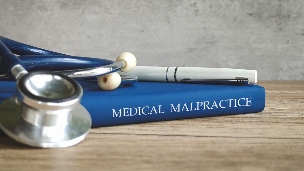 A blue medical law book titled "MEDICAL MALPRACTICE" on a wooden desk with a stethoscope and white pen for legal case documentation.
